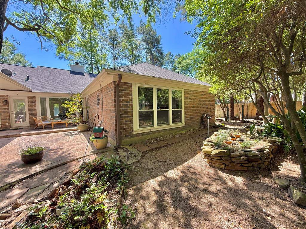 Charming backyard view featuring a brick exterior, large windows, and a cozy patio area. The space is surrounded by lush greenery, mature trees, and a stone-bordered planter, creating a serene outdoor retreat.