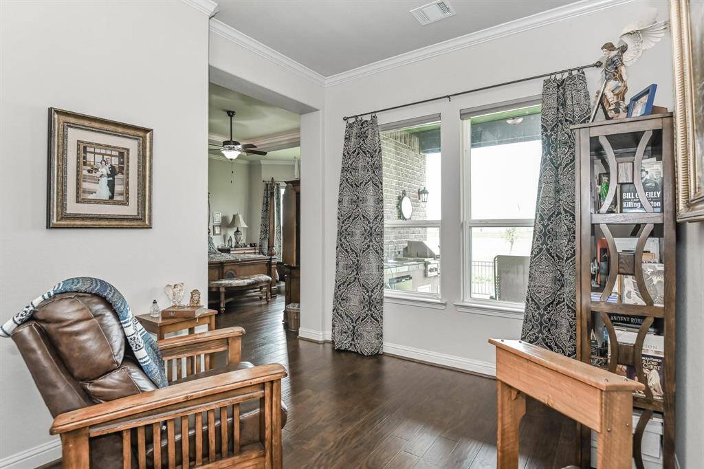 This cozy living space features elegant wood flooring, large windows with patterned curtains, and a comfortable leather armchair. A decorative shelf adds character, while the open layout connects to a room with ceiling fan and natural light, creating a welcoming atmosphere.