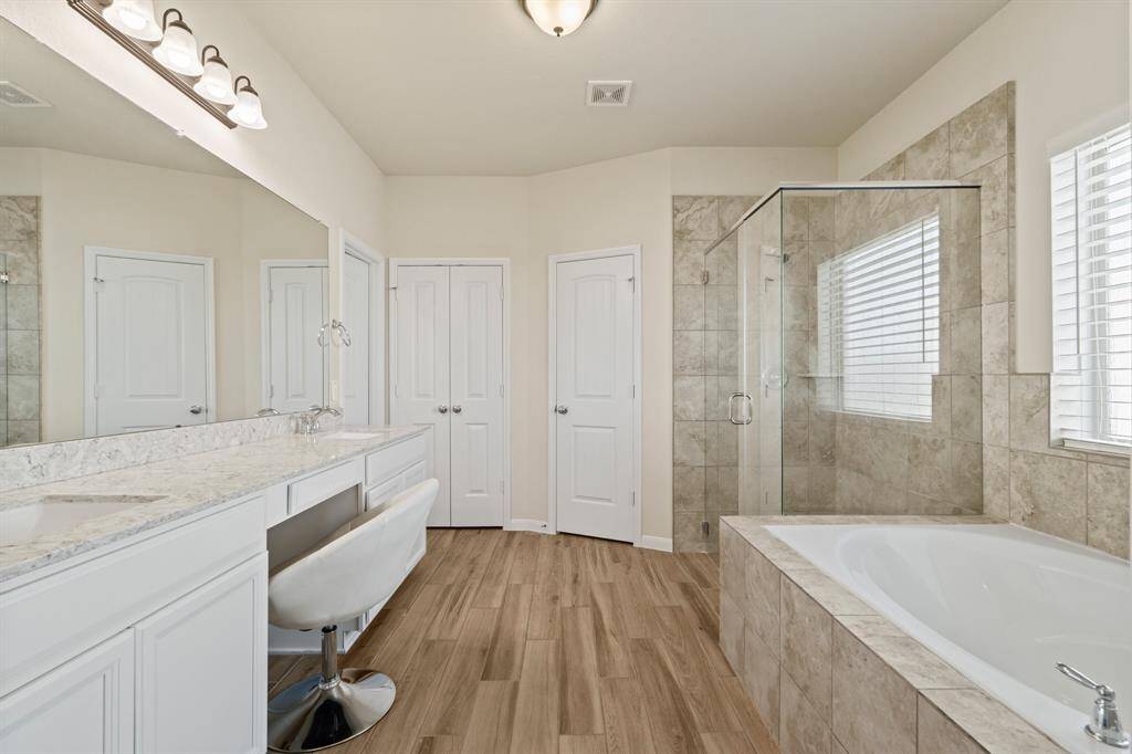 This spacious primary bathroom features a modern design with a large glass-enclosed shower, a separate soaking tub, and a double vanity with marble countertops. The light wood floors and ample natural light create a warm, inviting atmosphere.