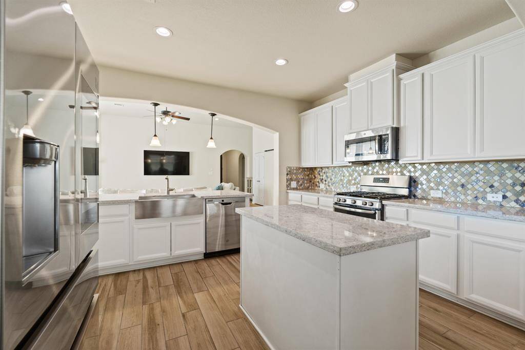 This modern kitchen features sleek white cabinetry, stainless steel appliances, and a stylish mosaic tile backsplash. The space is bright and open, with a central island and wood-look flooring, perfect for cooking and entertaining.
