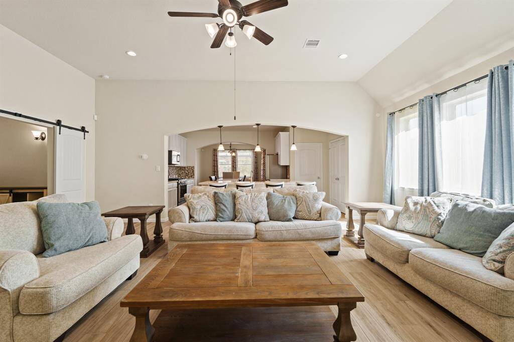 This inviting living room features high ceilings, a ceiling fan, and ample natural light from large windows with blue curtains. Opens seamlessly to a dining area and kitchen.