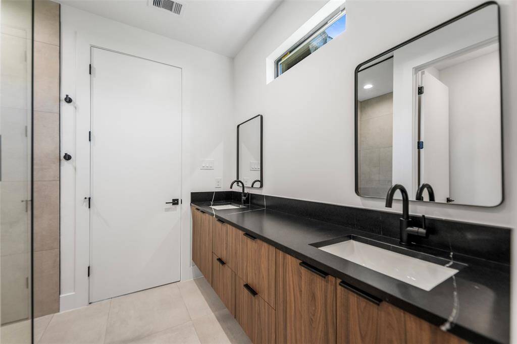 Modern en-suite bathroom featuring dual vanities with black quartz countertops, matte black fixtures, and custom wood cabinetry. Framed mirrors and a transom window add to the elegance.