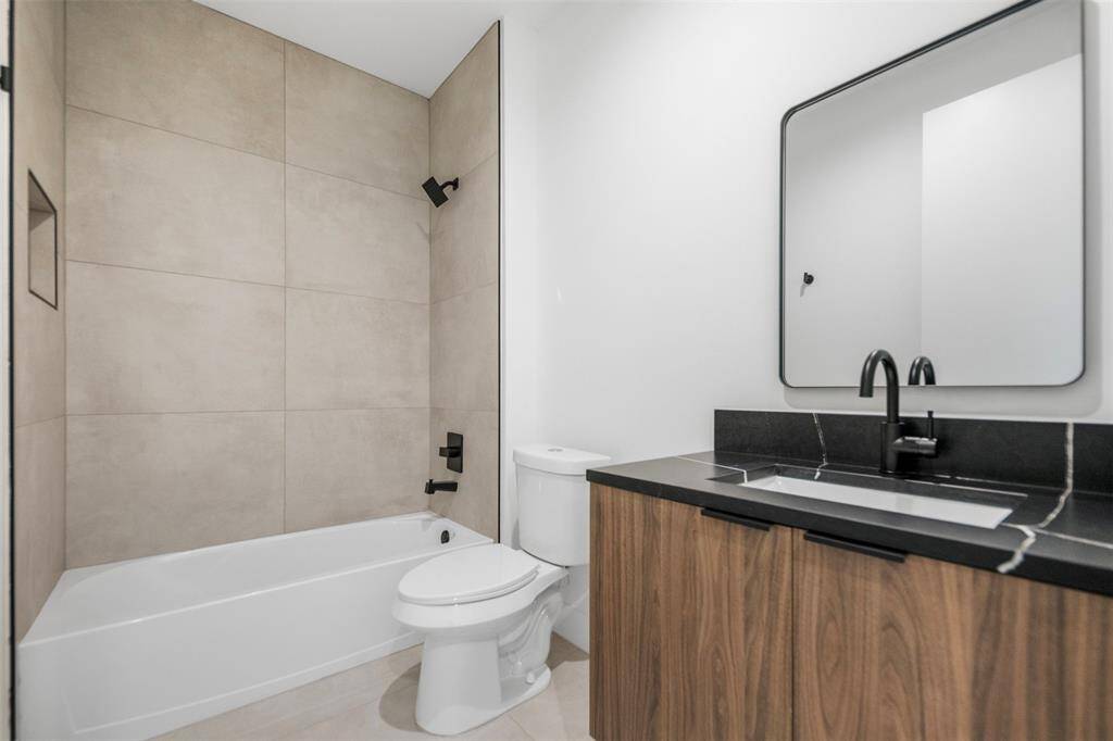 Contemporary full bath with an oversized tile tub surround, black hardware, and vanity with black quartz countertops and natural wood cabinetry