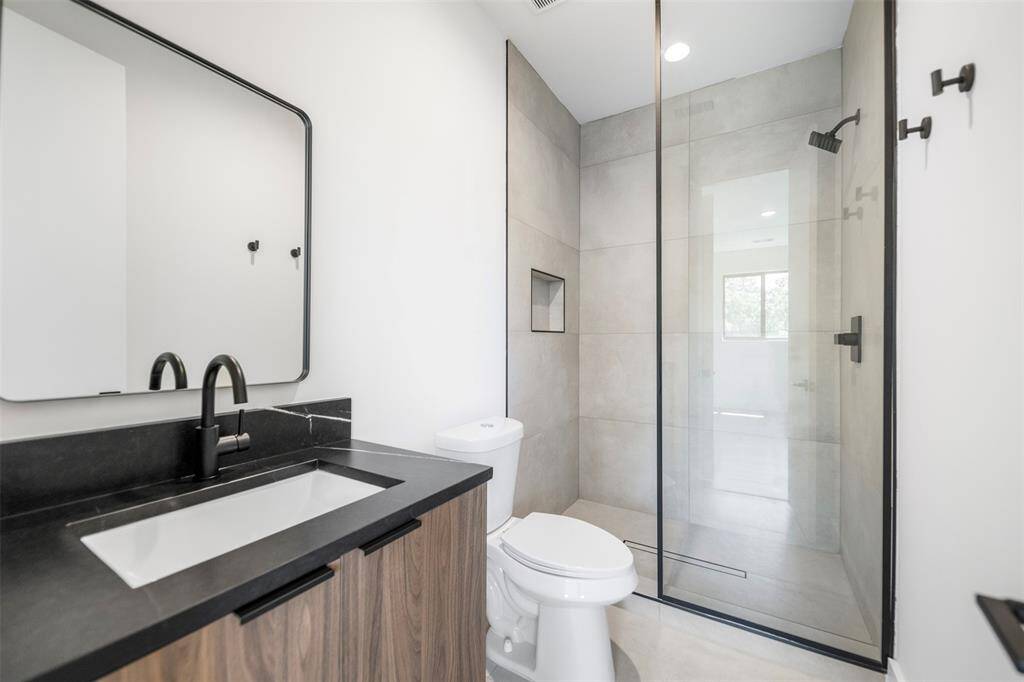 A sleek secondary bathroom featuring a walk-in shower with frameless glass, floor-to-ceiling tile, matte black fixtures, and a modern vanity with wood cabinetry and black quartz counters.