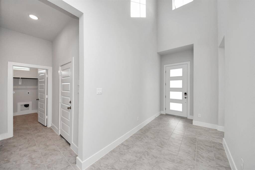 This photo shows a bright, modern entryway with high ceilings and tiled floors. It features a contemporary front door with frosted glass panels, and multiple doors leading to other rooms, including a utility area. The space is open and welcoming, ideal for creating a great first impression.