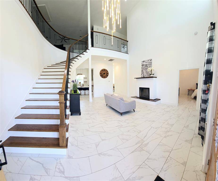 This entryway features a grand, curved staircase with elegant railings, high ceilings, and modern lighting. The room is bright with white walls and tile flooring, leading to a cozy living area with a sleek fireplace.