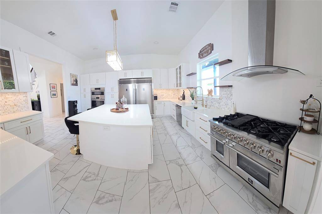 This spacious kitchen features modern white cabinetry, a large central island, and sleek stainless steel appliances, including a professional-grade range. The room is bright with ample natural light and elegant pendant lighting, complemented by quartz countertops, a stylish tile backsplash and marble-look flooring. Perfect for cooking and entertaining.