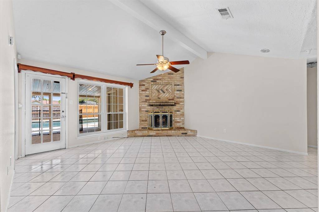 Spacious living room with high ceilings, a charming brick fireplace, and abundant natural light from large windows and French doors leading to the pool area. Perfect for relaxing and entertaining.