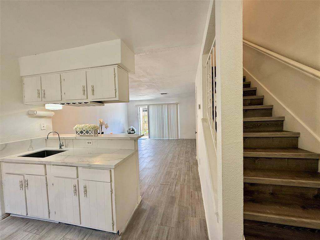 This photo showcases a bright, open-concept living space with a white kitchen featuring quartz countertops and ample cabinetry. There's a view of a living area with sliding doors leading to the outside, and a staircase with wooden steps.