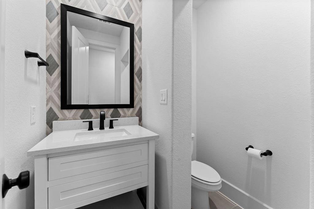 Half bath located off the utility room  features a sink with quartz counter top and a decorative tile wall, tile flooring, partitioned toilet area, and a framed mirror.
