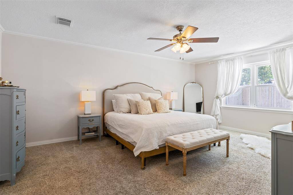 The master suite features a cozy, carpeted floor with neutral tones, a ceiling fan, and ample natural light from large windows creating a comfortable and inviting atmosphere.