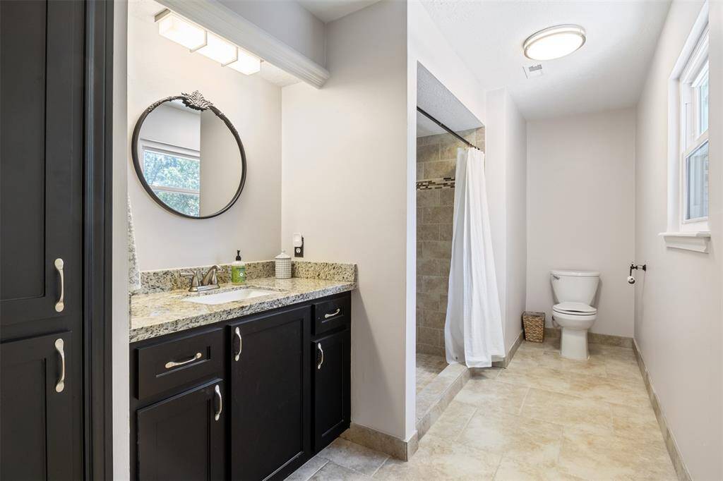 This bathroom features a modern design with a granite countertop, dark cabinetry, and a round mirror above the sink. It includes a spacious walk-in shower with a curtain and a separate toilet area. The room is well-lit with natural light from a window and overhead fixtures.