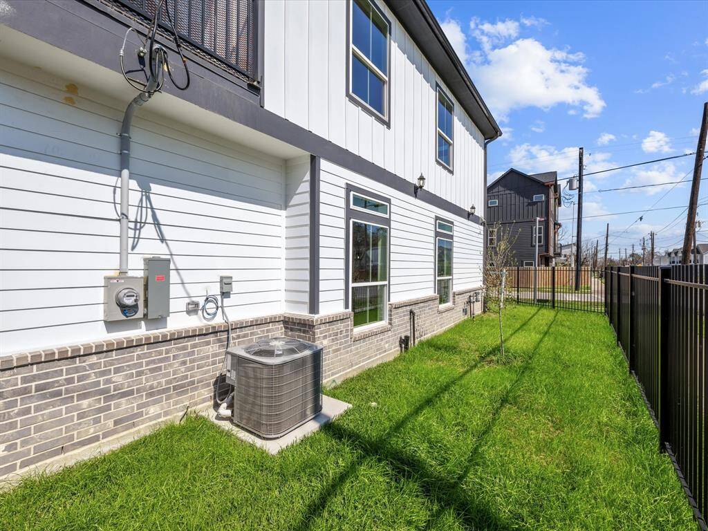 The photo shows a side view of a modern home with clean white siding and brick accents. It features large windows, a new air conditioning unit, and a neatly landscaped grassy yard enclosed by a black metal fence.