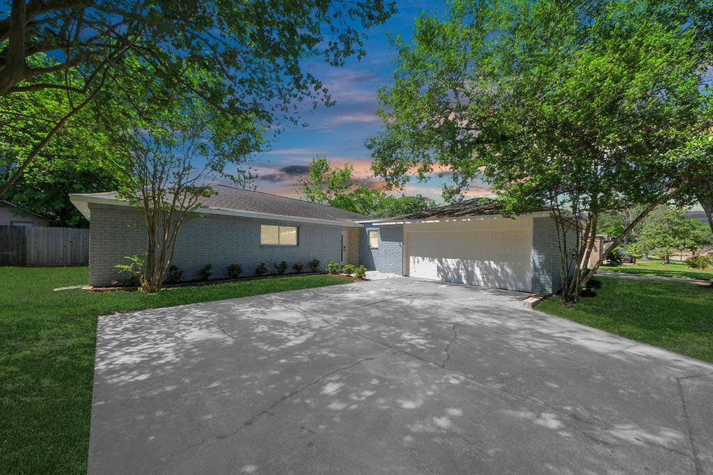 This photo shows a single-story home with a light-colored brick exterior and a two-car garage. It's surrounded by a well-maintained lawn and mature trees, creating a serene and inviting atmosphere. The driveway is spacious, and the sky above features a beautiful sunset.