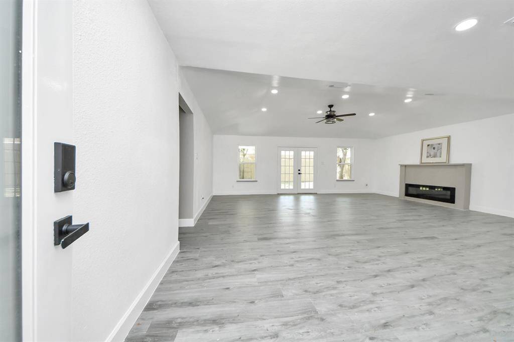 Spacious, modern living area with light gray flooring and white walls. Features recessed lighting, a ceiling fan, and a sleek fireplace. French doors and windows provide natural light and access to the outdoors.