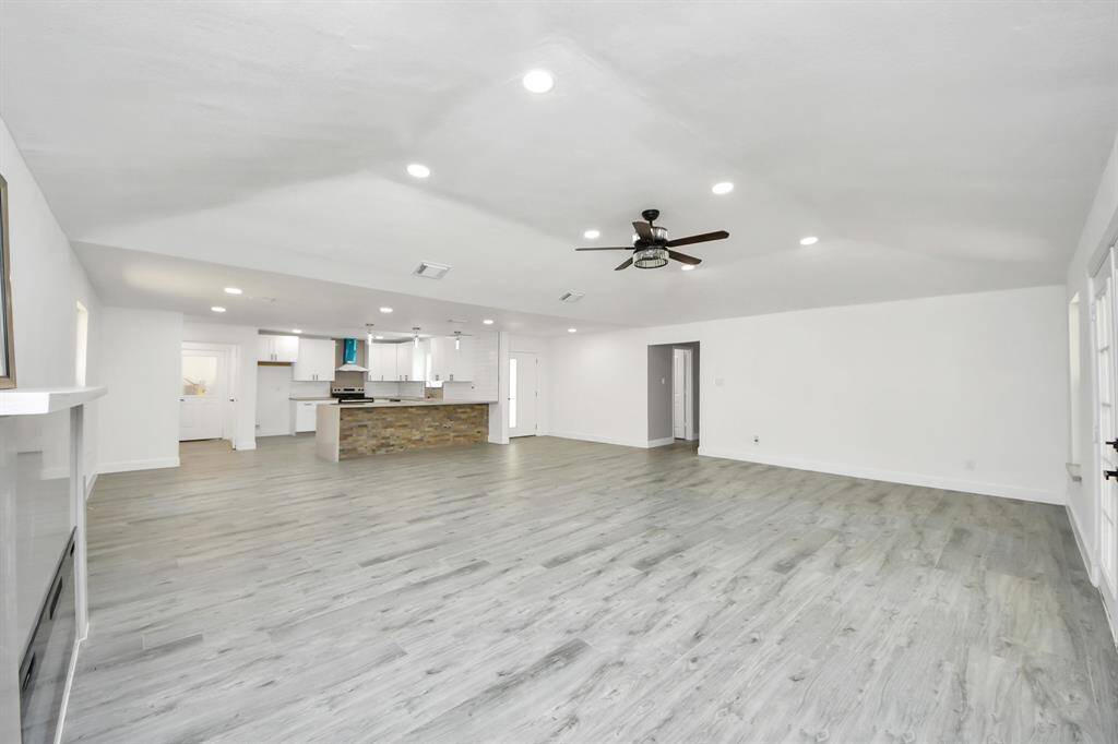 This spacious open-concept living area features modern light gray flooring, recessed lighting, and a ceiling fan. It seamlessly connects to a sleek kitchen with a stone-accented island, offering a bright and airy atmosphere ideal for entertaining.