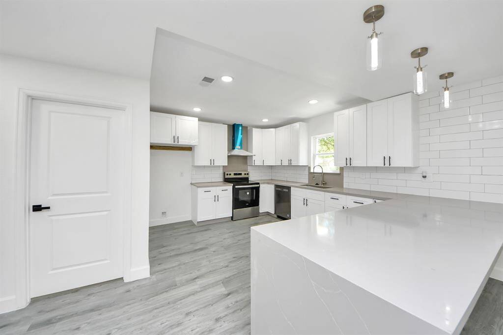 This modern kitchen features sleek white cabinetry, stainless steel appliances, and a spacious quartz countertop island. The room is brightened by recessed lighting and pendant lights, with a clean, open layout and stylish subway tile backsplash.
