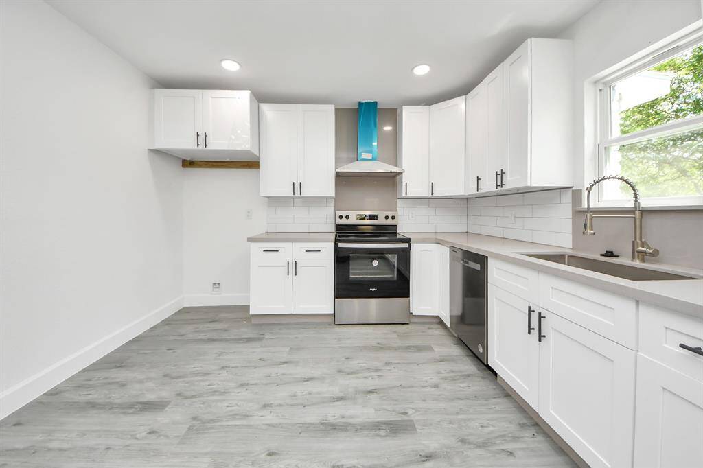 This modern kitchen features sleek white cabinetry, stainless steel appliances, and a spacious layout with ample counter space. Natural light pours in through a large window, highlighting the elegant backsplash and wood-style flooring.