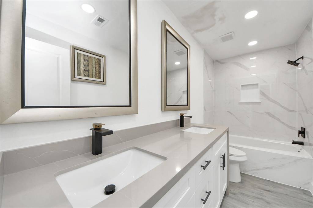 This bathroom features a modern double vanity with sleek black fixtures, elegant mirrors, and a spacious bathtub with marble-style tiles. The clean, neutral tones create a bright and welcoming atmosphere.