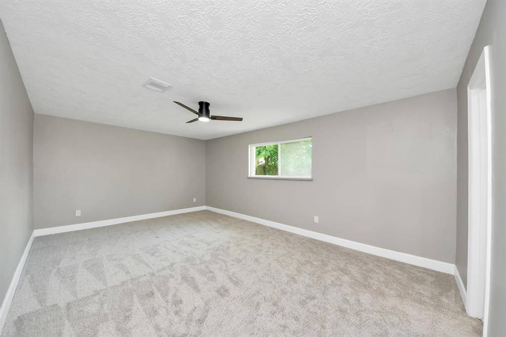 Spacious, carpeted room with neutral gray walls and a modern ceiling fan. A window provides natural light and a view of greenery outside.