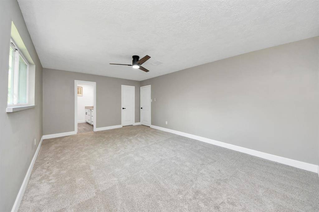 Spacious, carpeted bedroom with neutral walls, a ceiling fan, and natural light from a window. It leads to a bathroom and has two additional doors, likely for closets.
