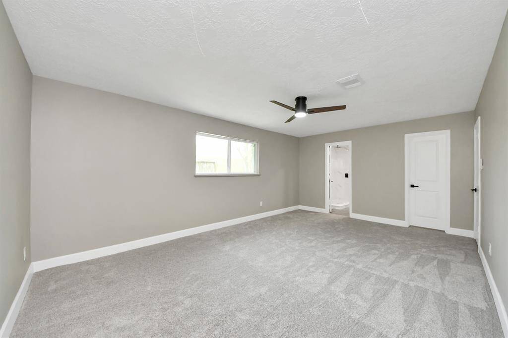 This spacious bedroom features soft gray carpeting and walls, a ceiling fan, and ample natural light from a large window. It includes an ensuite bathroom and a separate closet door, offering convenience and privacy.