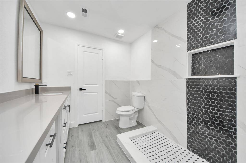 This modern bathroom features sleek gray flooring, a spacious vanity with ample storage, and a large mirror. The shower area showcases stylish black hexagonal tiles, complemented by elegant marble-patterned walls.