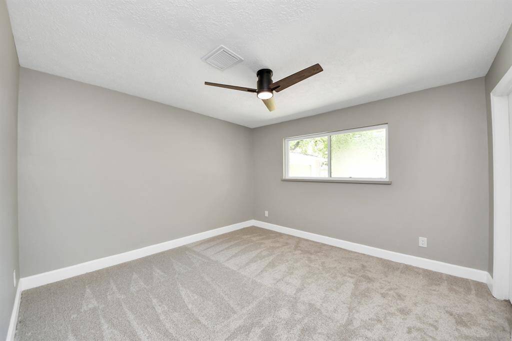 This photo shows a bright, unfurnished room with neutral gray walls, a modern ceiling fan, and a large window that lets in natural light. The room has fresh carpeting, creating a clean and inviting space.