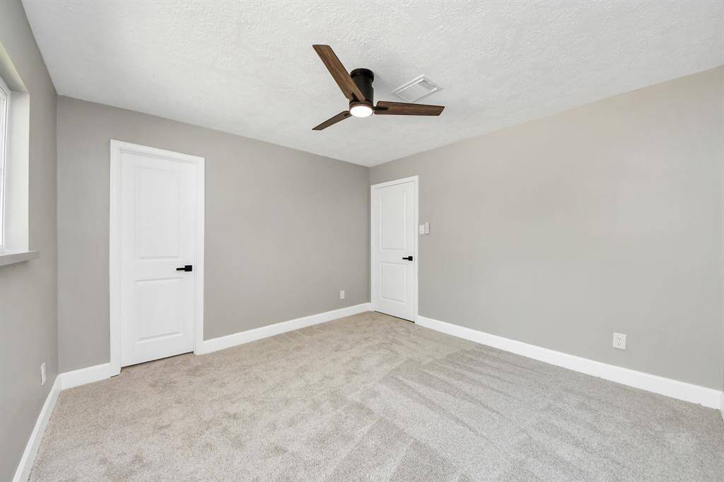 This room features neutral gray walls, plush carpet, and a modern ceiling fan. It has two white doors and a window for natural light, creating a bright and inviting space.