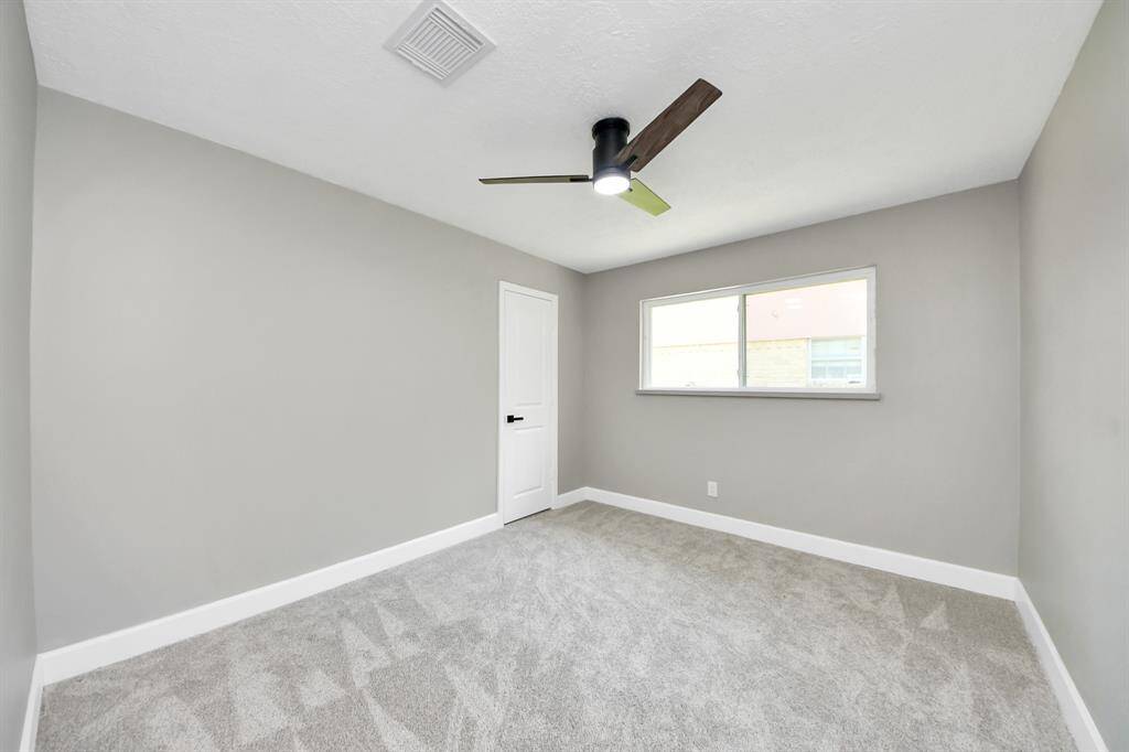 This room features neutral gray walls, a modern ceiling fan, and soft carpeting. A large window provides natural light, and a white door adds a clean touch, making it a versatile space.