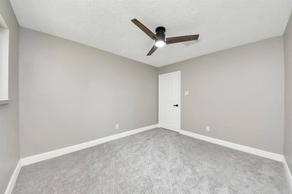 This room features soft gray walls, plush carpet, a modern ceiling fan, and a white door, offering a clean and contemporary feel.
