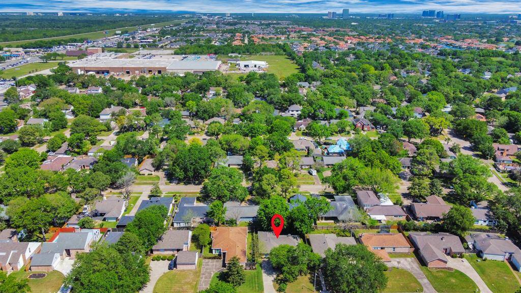 Aerial view of a suburban neighborhood with a mix of single-story homes surrounded by lush green trees. The area appears peaceful, with well-maintained properties and a nearby shopping center.