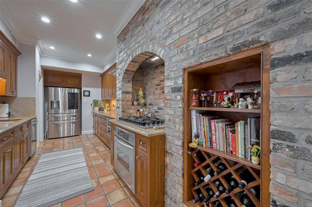 This kitchen features warm wood cabinetry, a stainless-steel refrigerator, and a brick arch housing a cooktop. There’s a built-in shelf with wine storage and books, and terracotta tile flooring adds rustic charm.