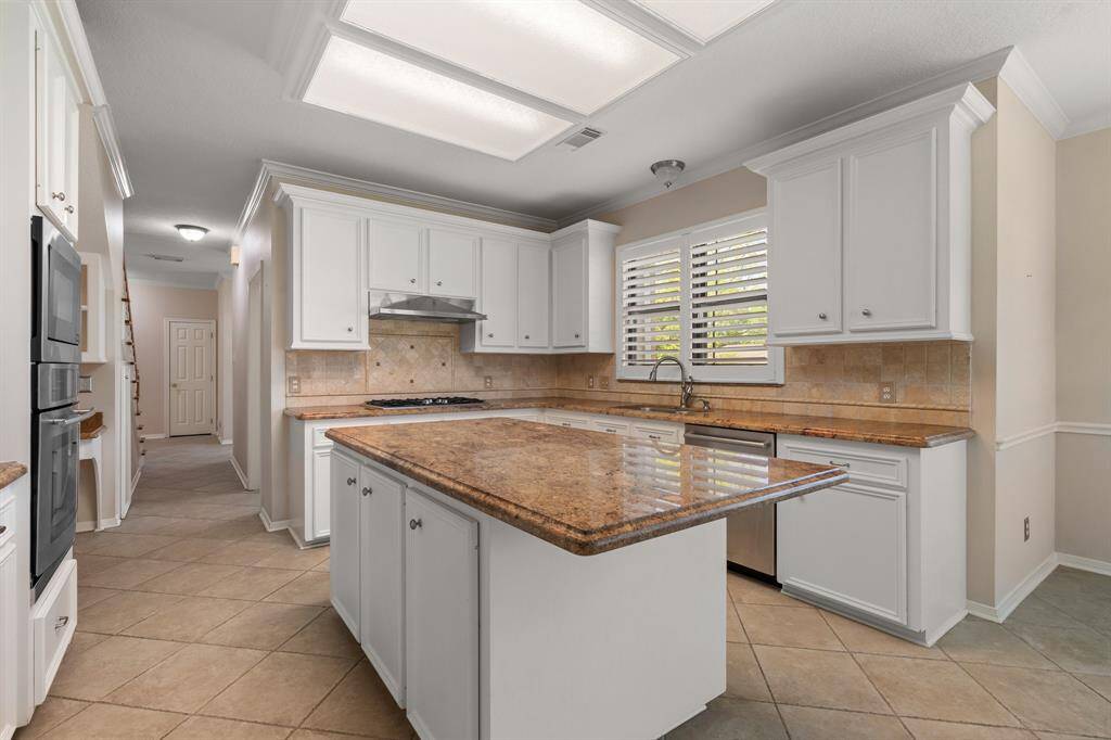This spacious kitchen features modern white cabinetry, granite countertops, and a central island. It includes stainless steel appliances, tile flooring, and ample natural light from large windows with shutters.