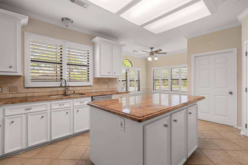 This bright kitchen features white cabinetry, a large granite island, and ample natural light through plantation shutters. The tile flooring and ceiling fan add to its charm and functionality.