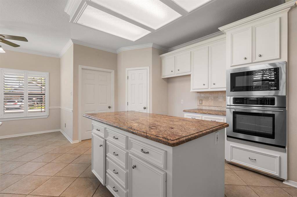 White cabinetry and a large central island featuring a brown granite countertop. It includes modern stainless steel appliances, ample storage, and a window with plantation shutters for natural light.