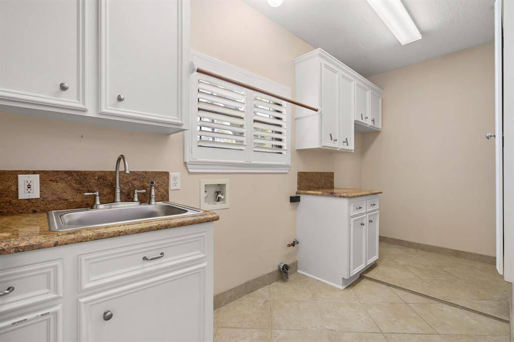 Laundry room showcases a stainless steel sink, and stone countertops. There's space for appliances and a window with shutters for natural light. The room has tiled flooring and ample storage.