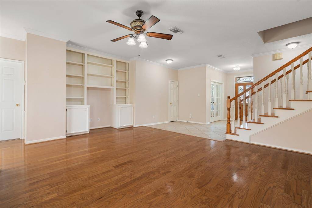 Living area features hardwood floors, built-in shelving, and a ceiling fan. The room is well-lit with neutral walls and has an elegant staircase leading to the upper level, creating a warm and welcoming atmosphere.