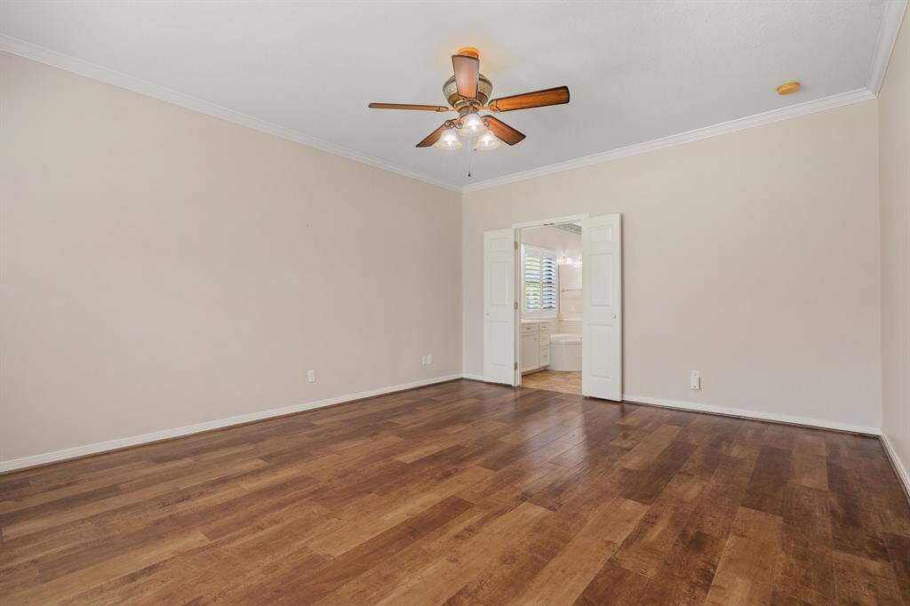 Primary bedroom down, features warm wood flooring, neutral walls, and a ceiling fan. It opens into a bright bathroom with double doors, offering a welcoming and functional layout.