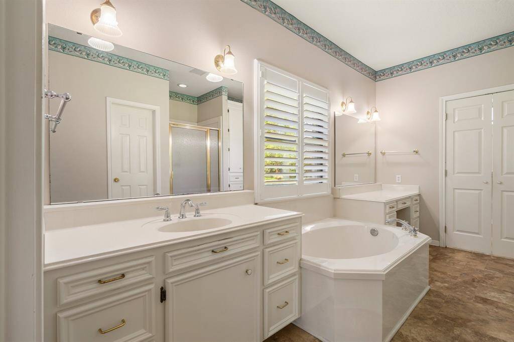 This bathroom features a spacious layout with a large soaking tub, double vanity with ample storage, and a separate shower. It has elegant lighting fixtures, a decorative border, and shuttered windows for natural light.