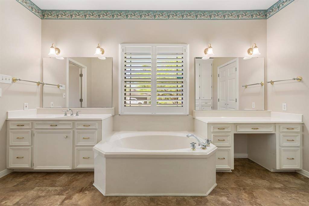Primary bathroom has a central soaking tub, dual vanities with ample storage, and a large window with shutters for natural light. The neutral color palette and decorative border is waiting for your personal touches!
