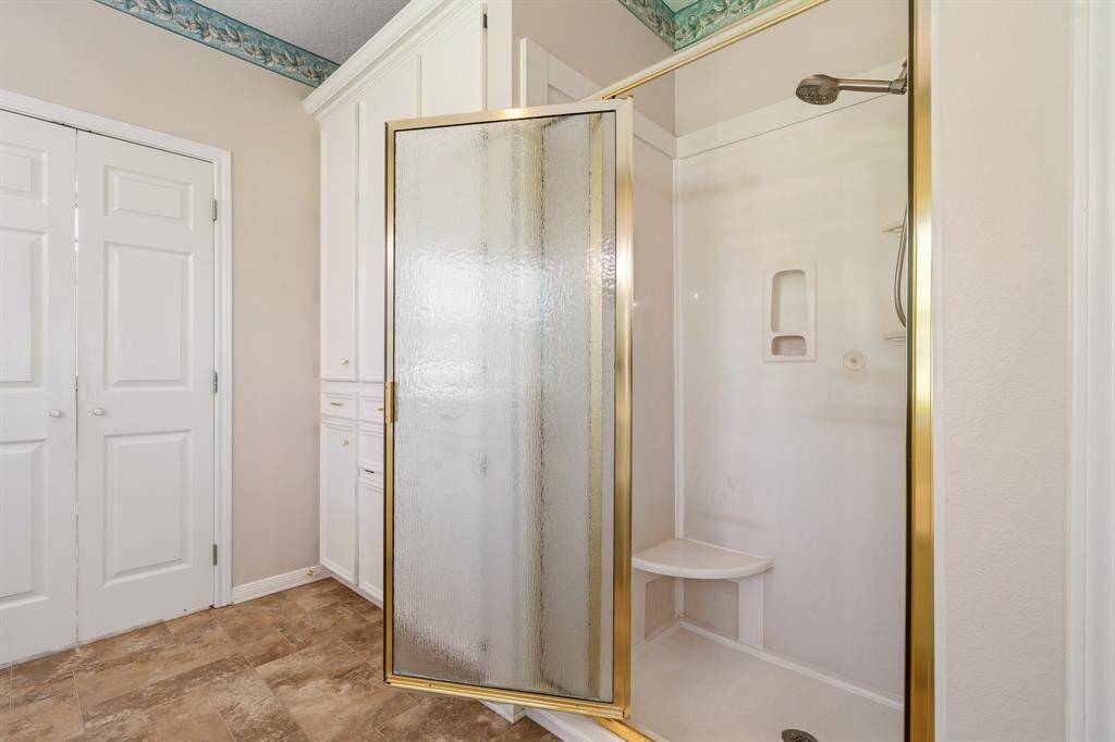 Primary bathroom features a shower with a frosted glass door and gold trim, built-in shelving, and a corner seat. The space includes ample storage with white cabinetry and has a neutral tile floor.