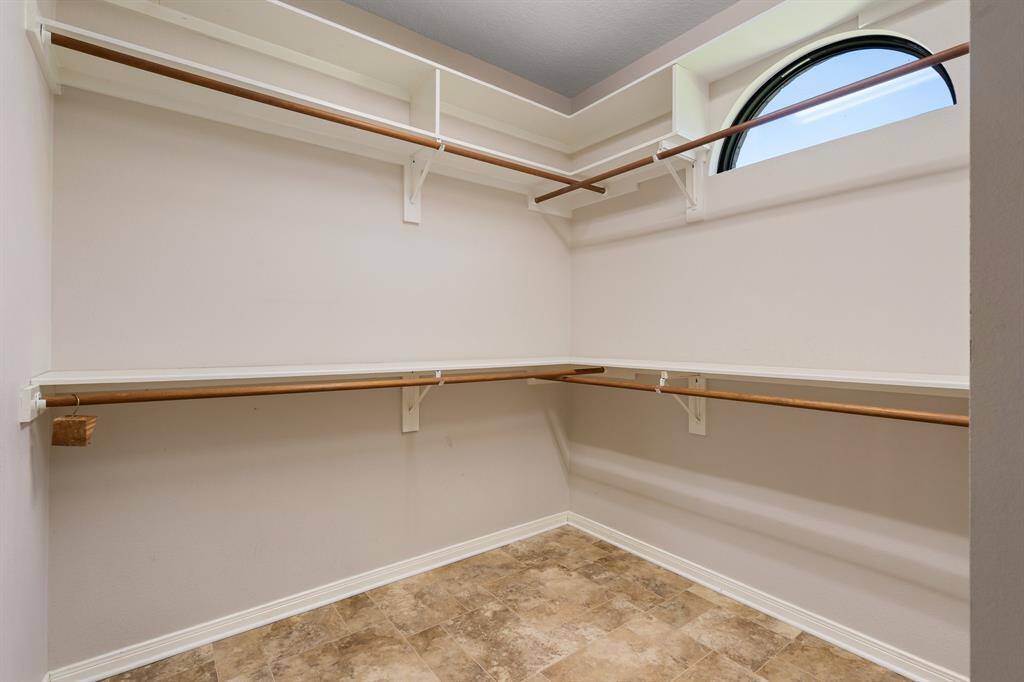 Primary walk-in closet features tons of hanging space with wooden rods, a high window for natural light, and a neutral tile floor, making it both functional and bright.