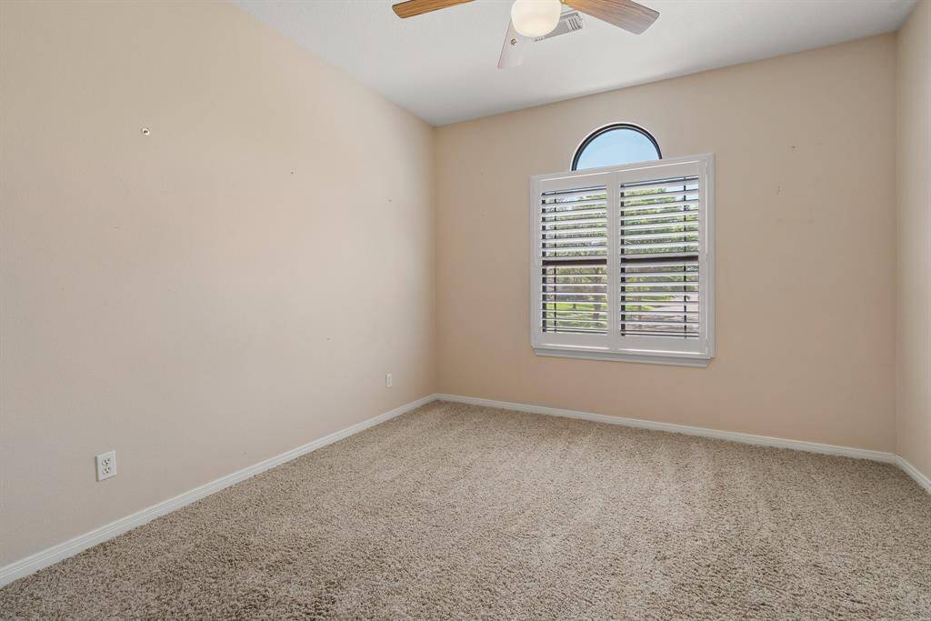 Third bedroom upstairs features neutral walls, plush carpeting, and a ceiling fan. A window with shutters provides natural light and a pleasant view. Perfect for a cozy bedroom.