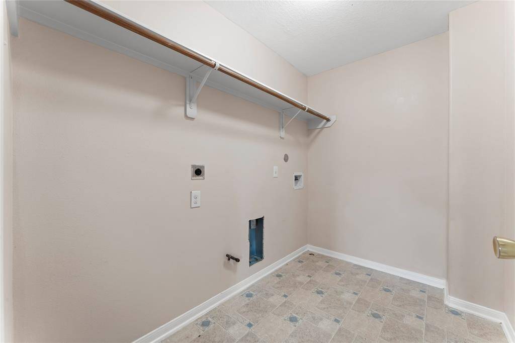 This photo shows a laundry room with hook-ups for a washer and dryer, beige walls, and a tiled floor. There's a wooden shelf and hanging rod for additional storage.