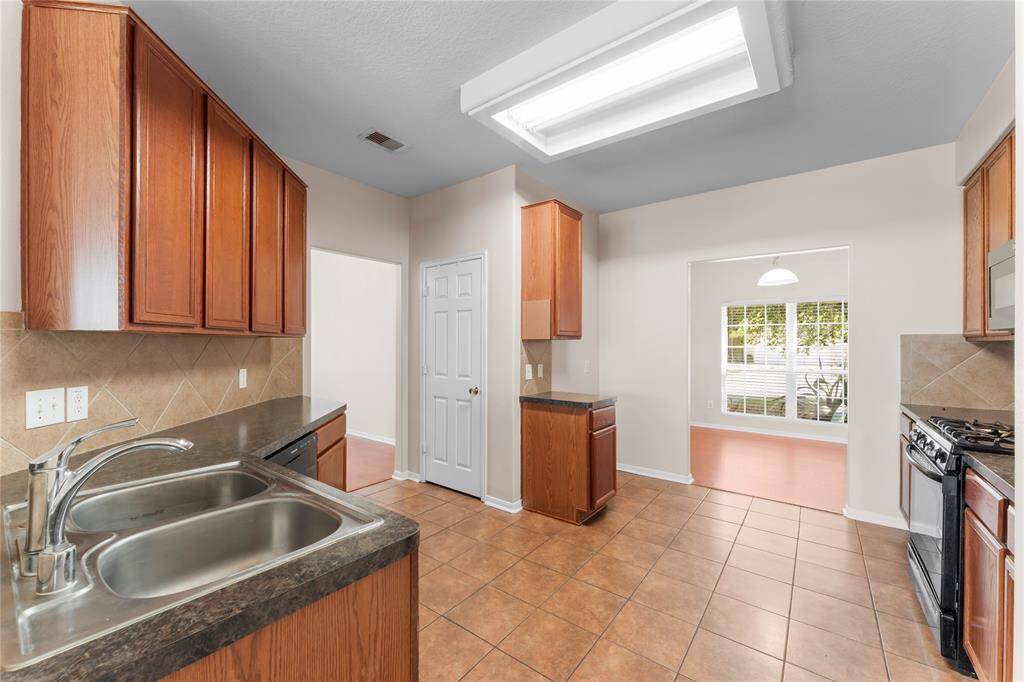 The kitchen features warm wood cabinets and dark countertops, complemented by a double sink. The space is brightened by a large window and overhead lighting, with tile flooring and a neutral palette.
