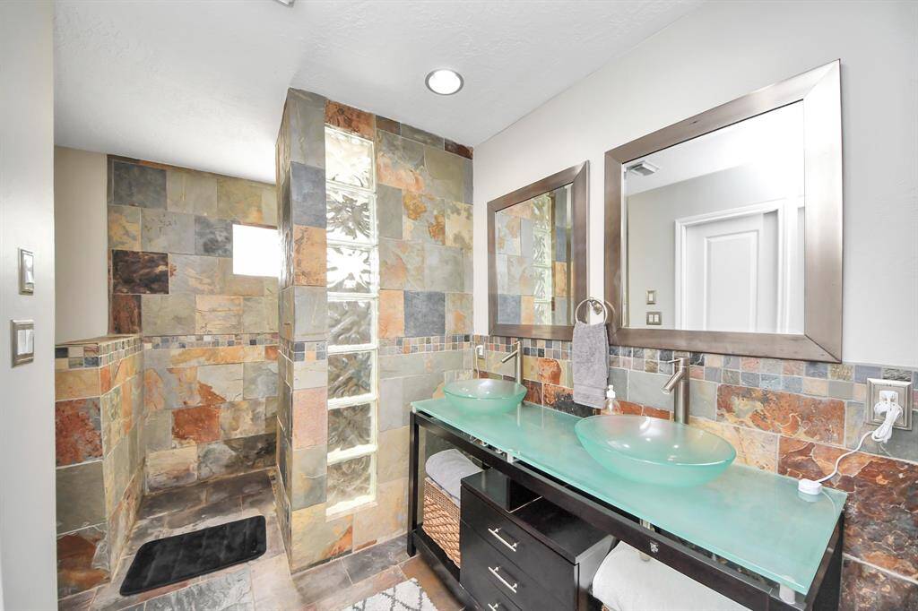 The modern bathroom features sleek above-bowl glass sinks and sleek cabinetry, creating a contemporary and refined atmosphere. The space is further enhanced by the elegant slate floors and a matching slate-tiled walk-in shower with block glass accent, offering a sophisticated and cohesive design.