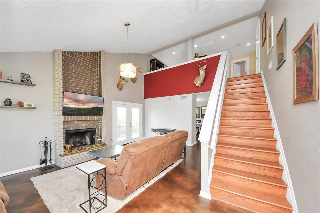 Soaring ceilings, a striking floor-to-ceiling brick fireplace, and sleek stained concrete floors create a warm yet modern living space. Natural light flows through the double glass doors, while the rich wood staircase and bold accent wall add character — simply perfect.