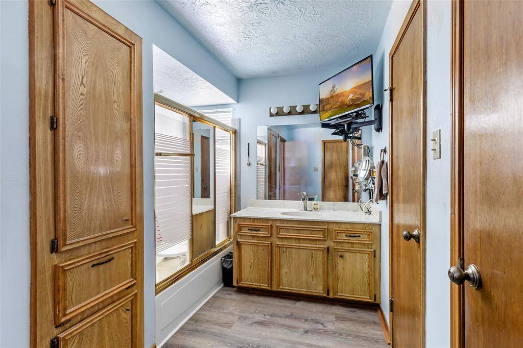 This primary bathroom features a spacious vanity with a large mirror, wood cabinets, and modern flooring. The tub/shower area is enclosed with frosted glass doors.