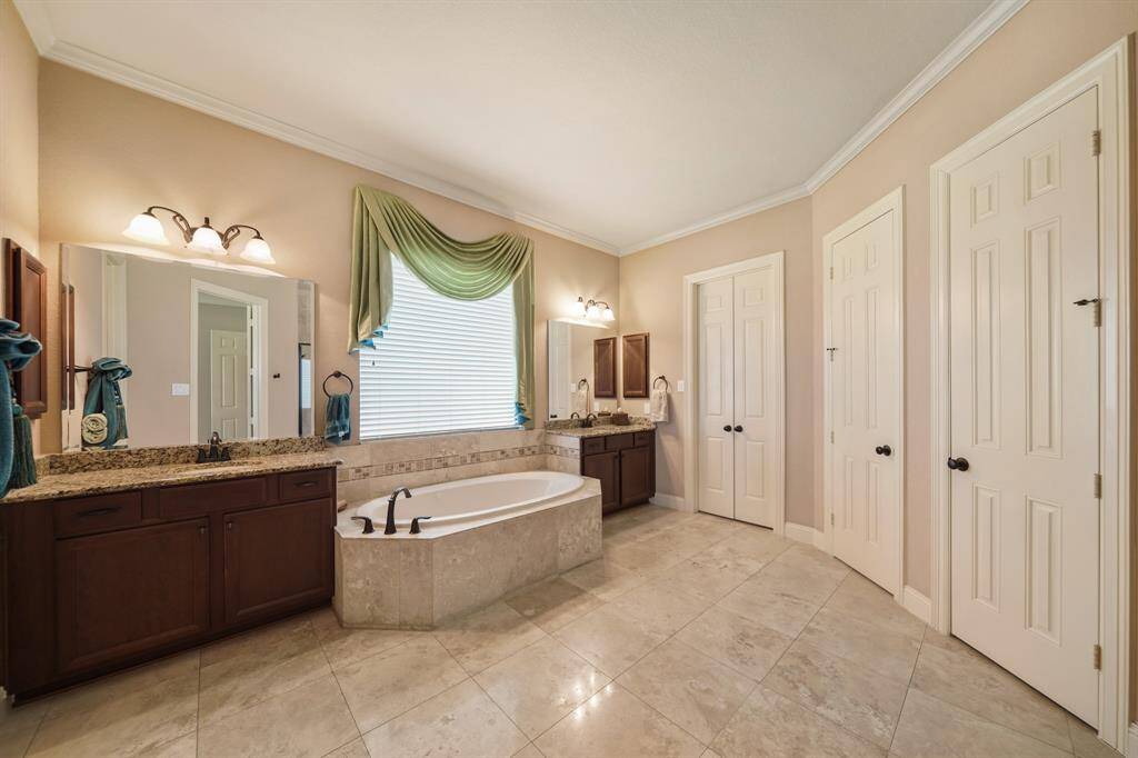 This spacious bathroom features a luxurious soaking tub, dual vanity with granite countertops, elegant lighting, and ample storage with multiple closets. The large window provides natural light, complementing the serene, neutral color palette.